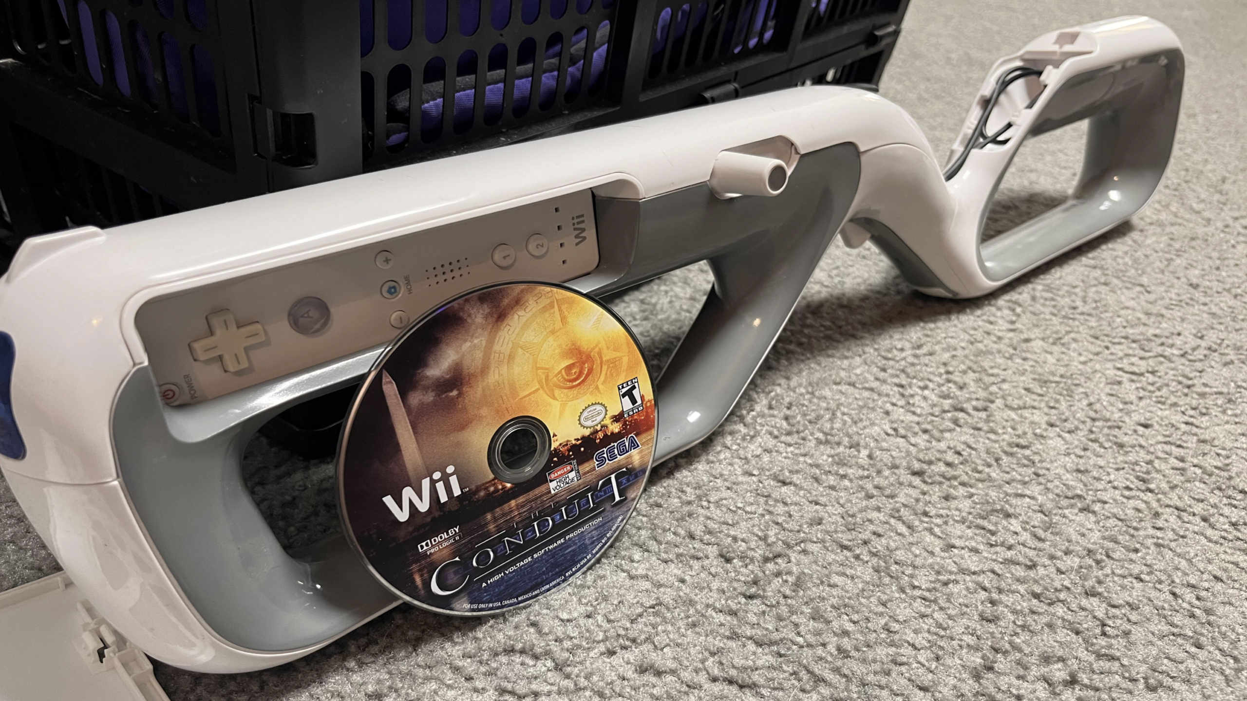 A Nintendo Wii controller and a white Wii zapper accessory, both essential gaming accessories, rest on a carpet, with a Conduit 2 Wii game disc partially inserted into the zapper.