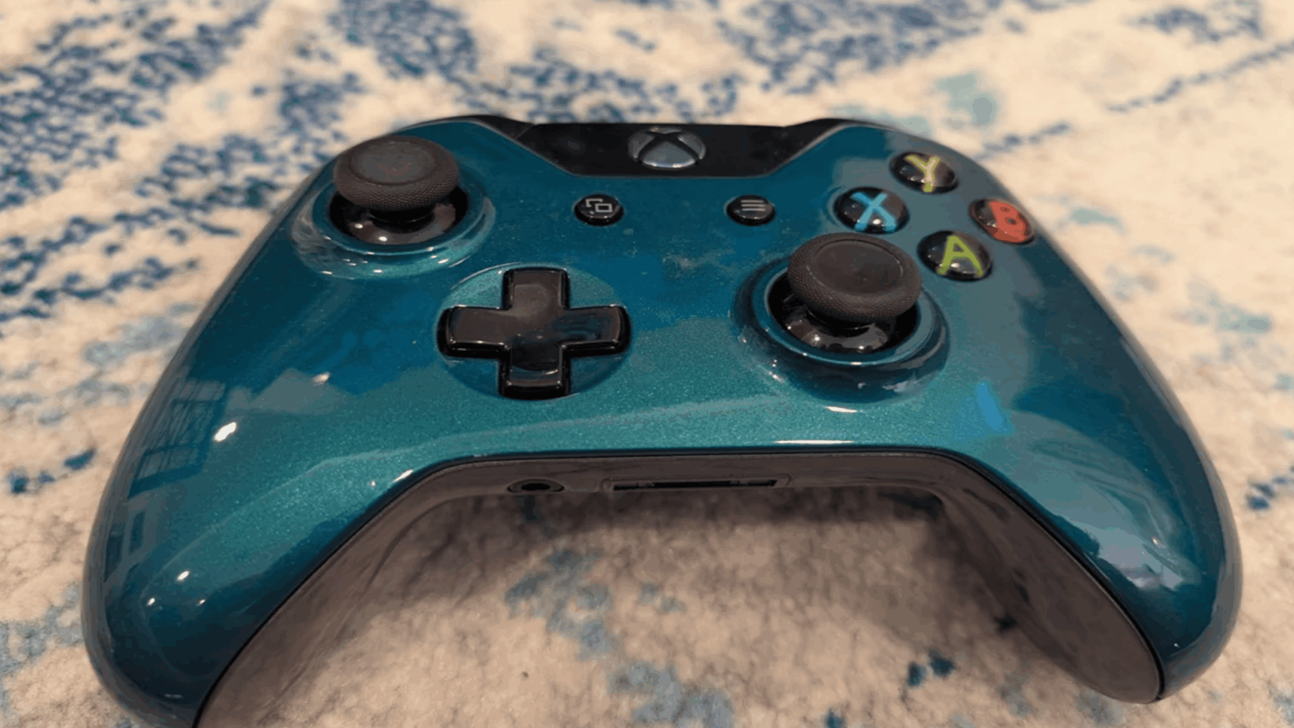 A shiny teal Xbox controller with black buttons and joysticks, a true symbol of expensive gaming, rests on a blue and white textured surface.
