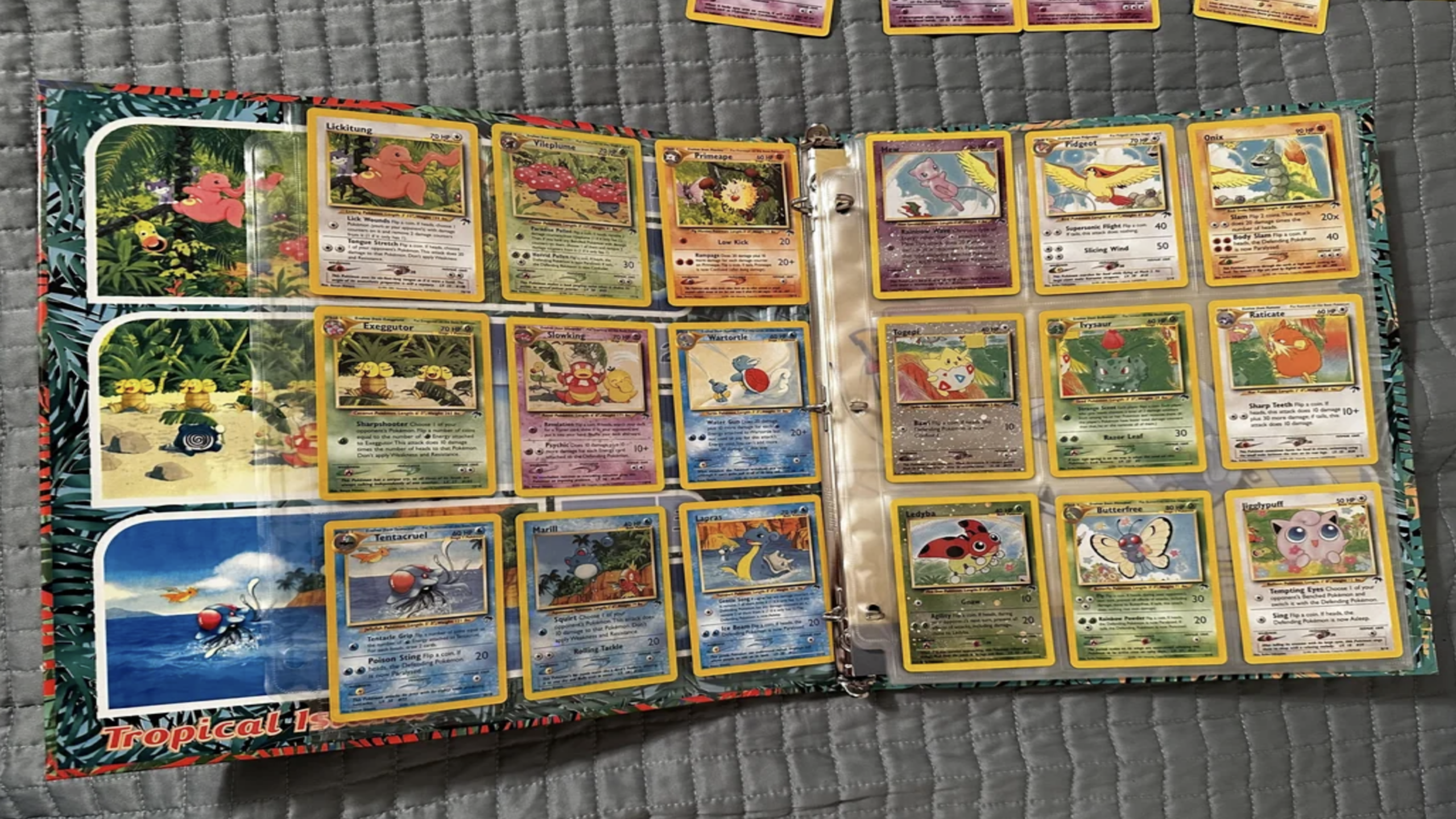 A binder open to two pages showing a collection of Pokémon trading cards displayed in plastic sleeves, all laid out on a gray quilted bedspread—a nostalgic scene filled with cherished childhood items.