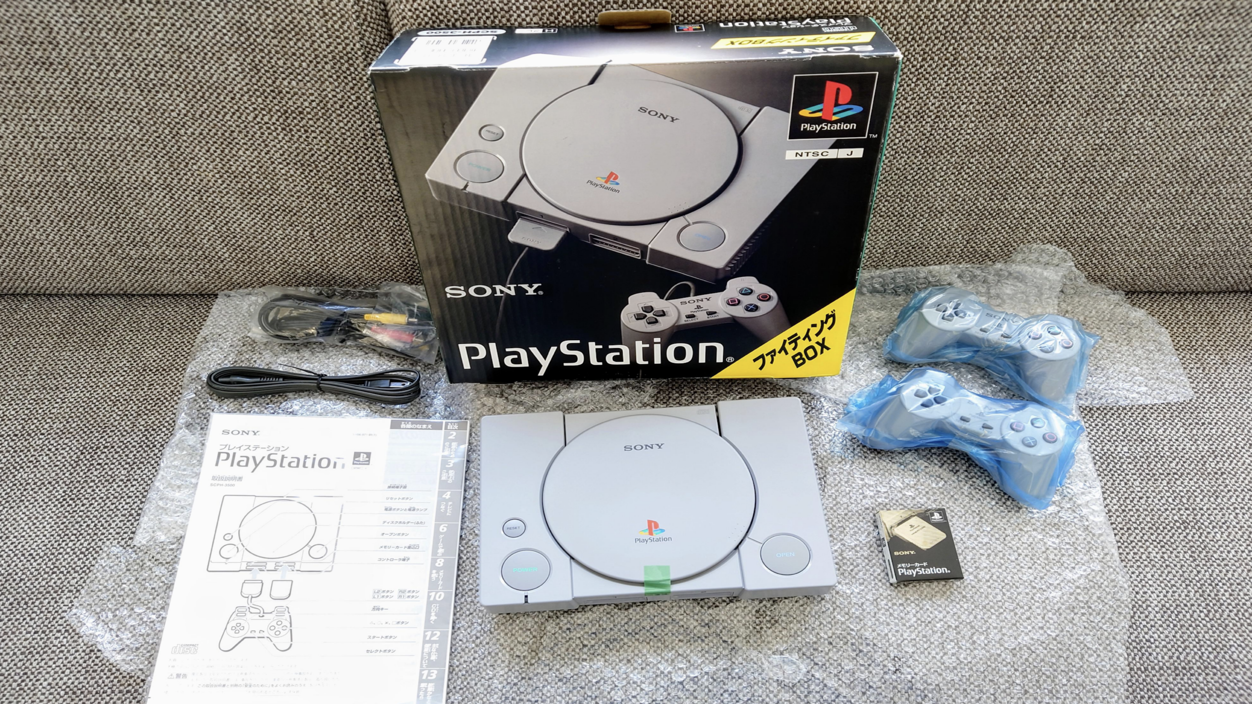Original Sony PlayStation console with box, two PlayStation controllers, instruction manual, and cables displayed on a beige surface with bubble wrap.