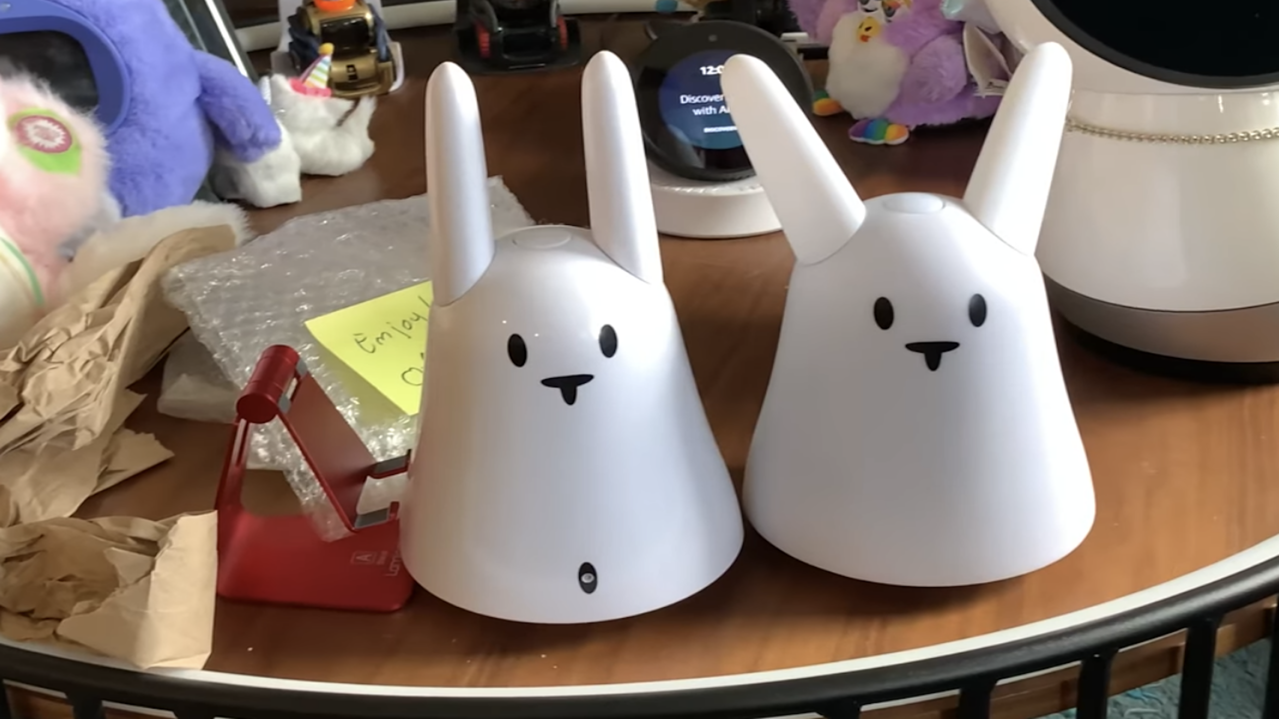 Two white, rabbit-shaped electronic devices with simple faces sit on a cluttered wooden table, surrounded by paper, a red phone stand, a smart display, an assortment of tech gadgets, and various toys.