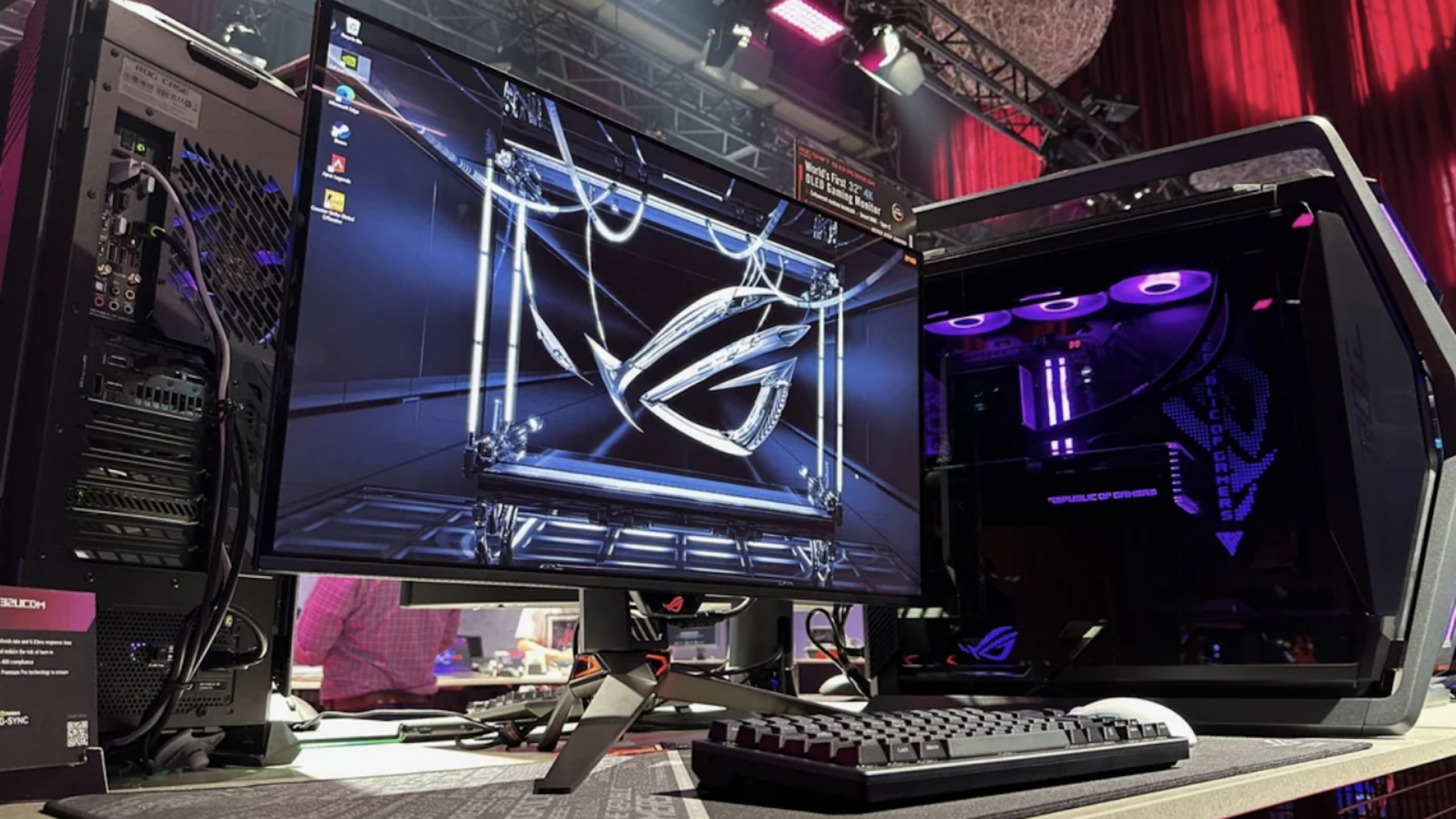 A gaming desktop setup featuring a large monitor displaying the ROG logo, a mechanical keyboard, and a PC with purple-lit fans—all premium Gaming Products—arranged on a desk in an event setting with red curtains and stage lighting.