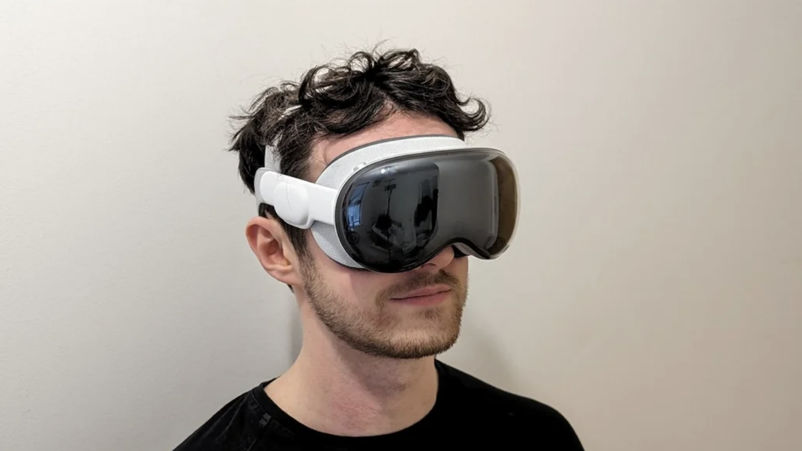 A person with short, curly dark hair wears a large, white and gray VR headset—one of the latest gaming products—standing against a plain, light-colored wall.