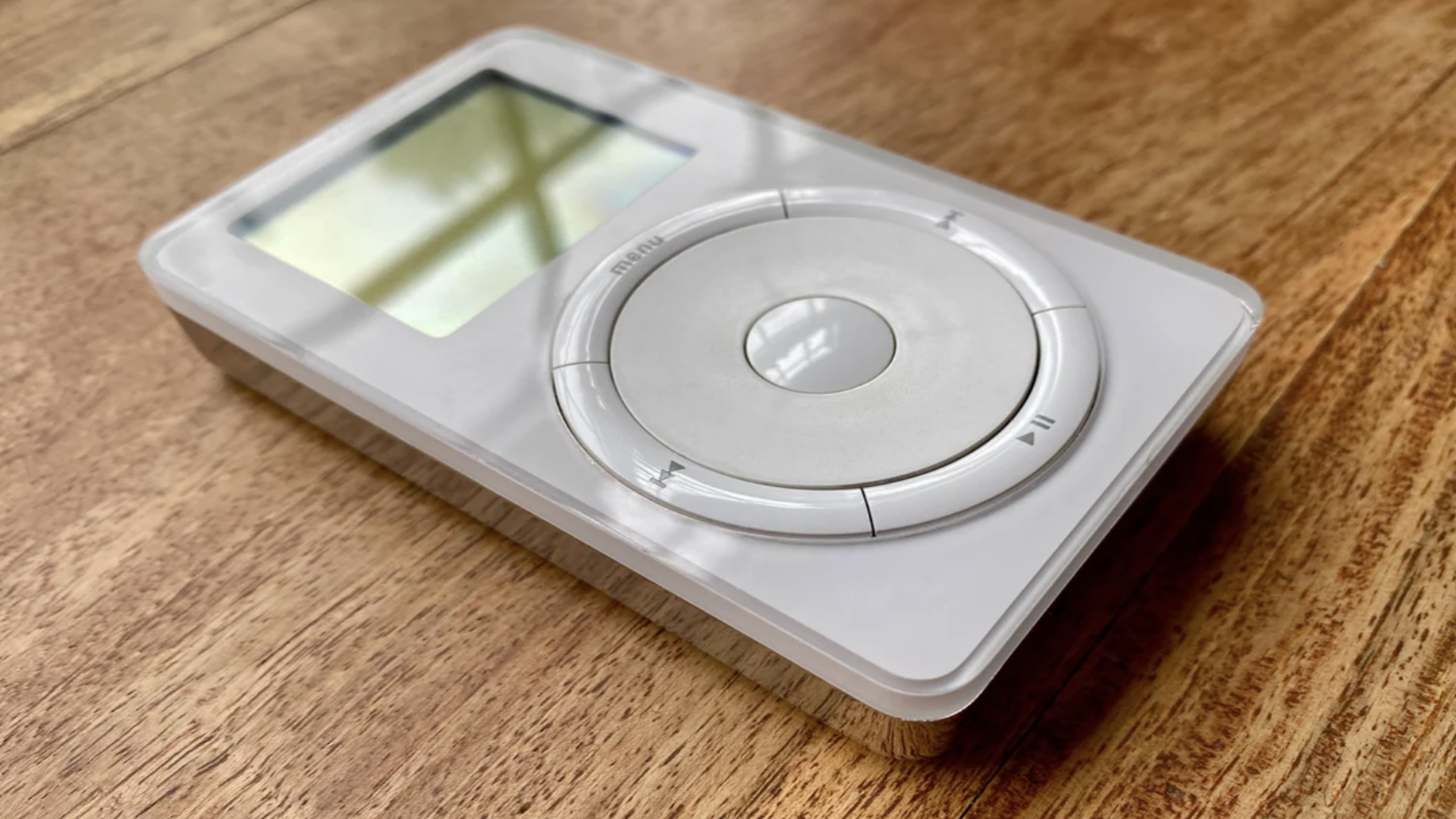 A first-generation white iPod with a circular scroll wheel and a small screen, one of those classic childhood items, resting on a wooden surface.