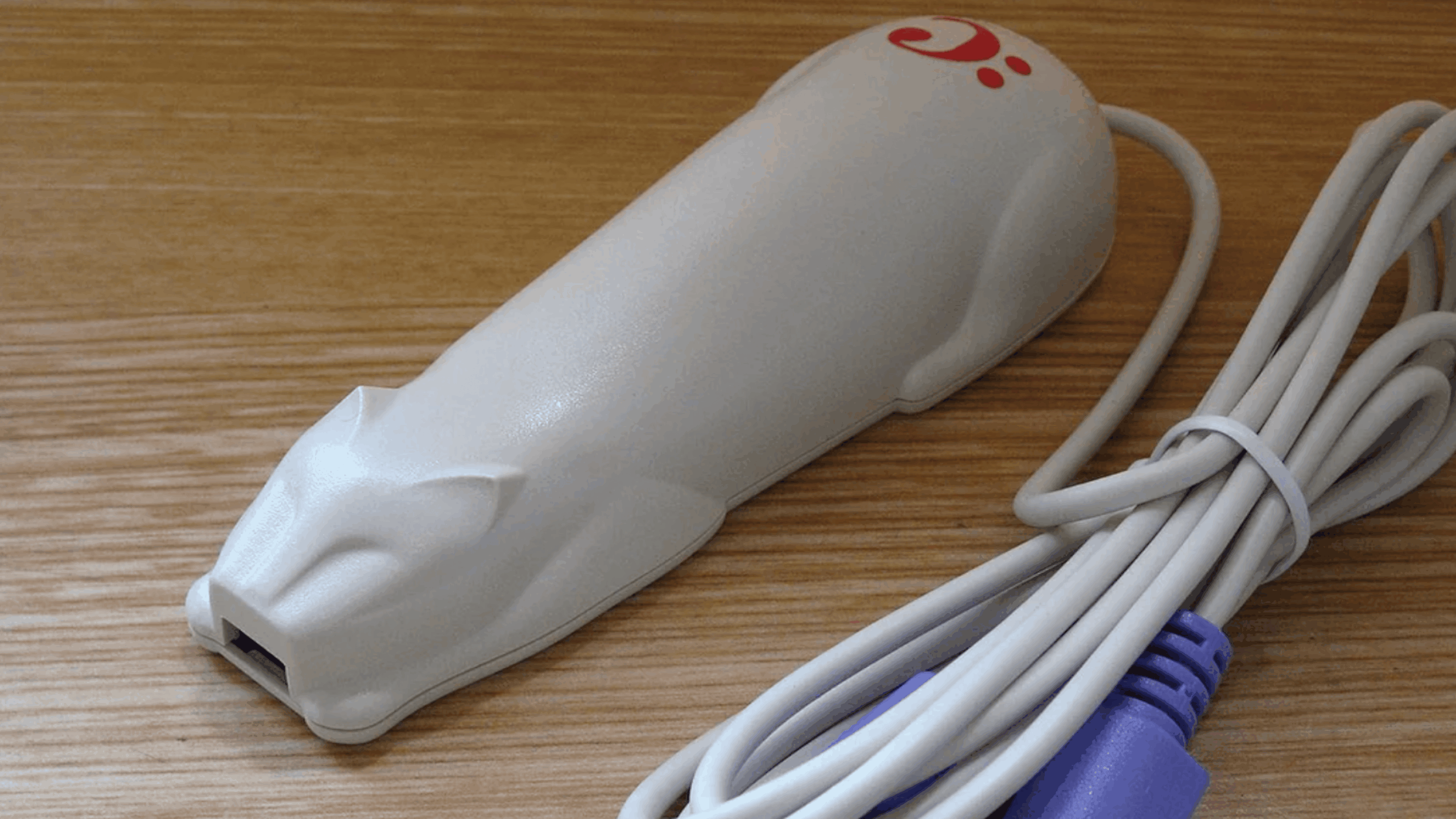 A computer mouse shaped like a white cat with a long cord, lying on a wooden surface. The cat shape is stylized and features a red logo near the tail, making it a charming addition for anyone who loves unique tech gadgets.