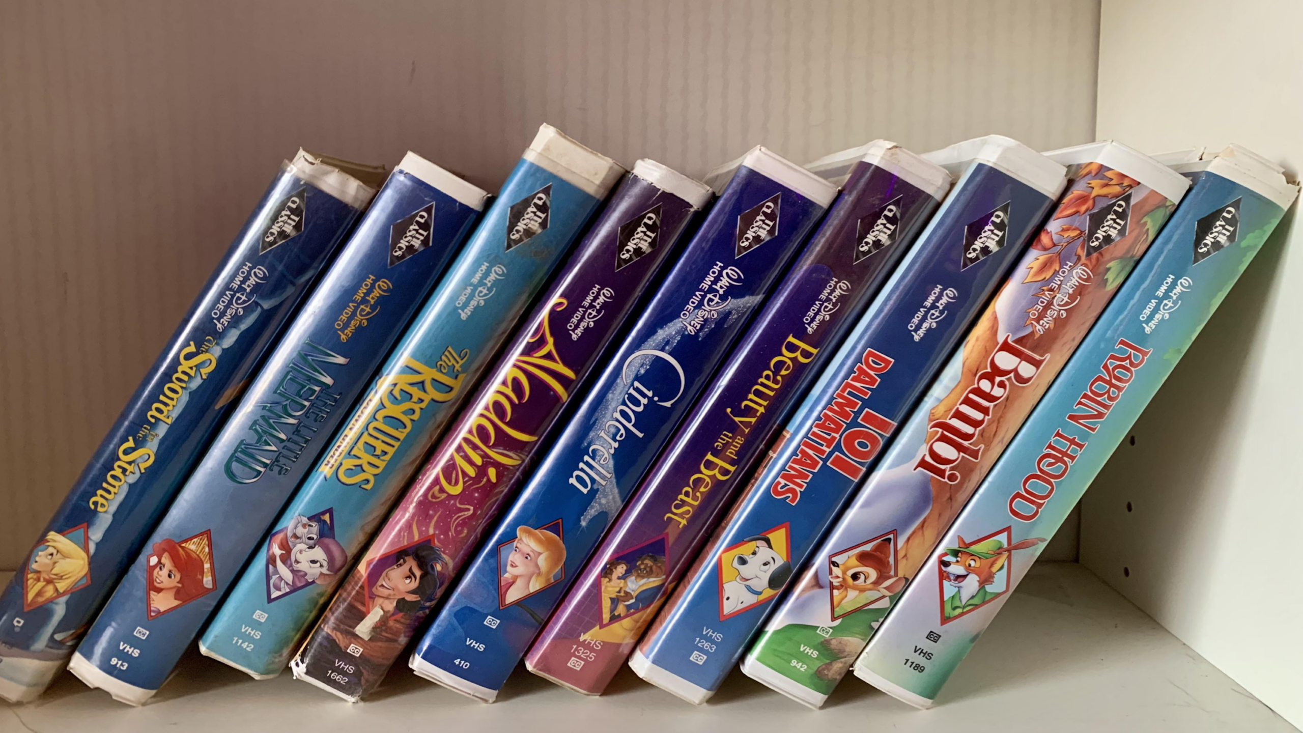 Nine Disney VHS tapes, cherished childhood items, including The Little Mermaid, The Rescuers, Aladdin, Cinderella, Beauty and the Beast, 101 Dalmatians, Bambi, and Robin Hood, are lined up on a white shelf, some leaning to the right.