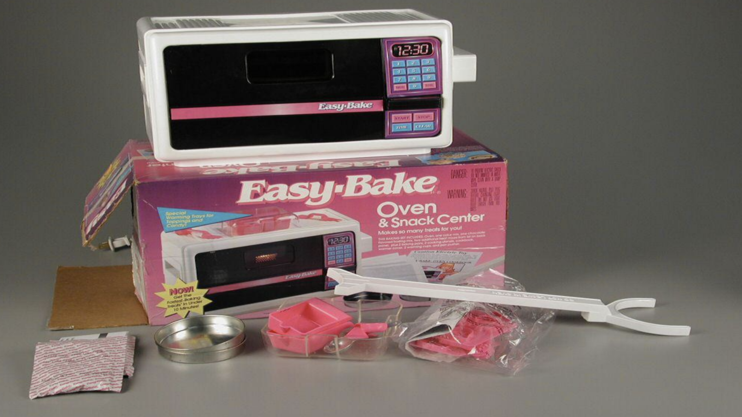 An Easy-Bake Oven and Snack Center with its box, baking accessories, pans, packets, and utensils—classic Childhood Items—are displayed on a plain background.