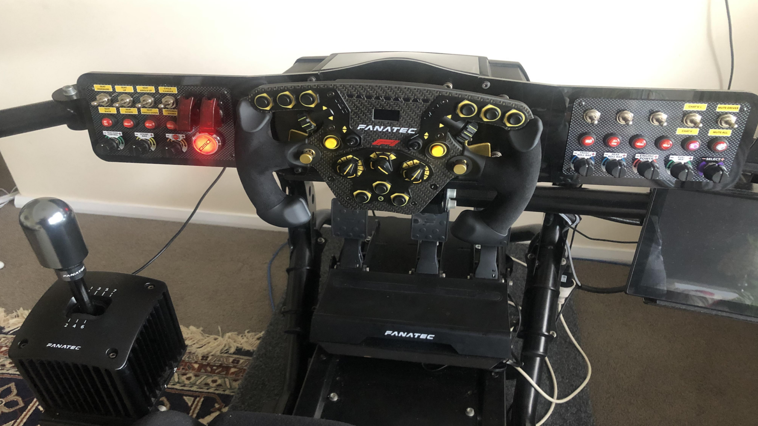 A high-end racing simulator setup featuring premium Gaming Products, including a Fanatec steering wheel, pedals, gear shifter, and button panels—all mounted securely on a sleek frame, with cables and a small screen visible on one side.