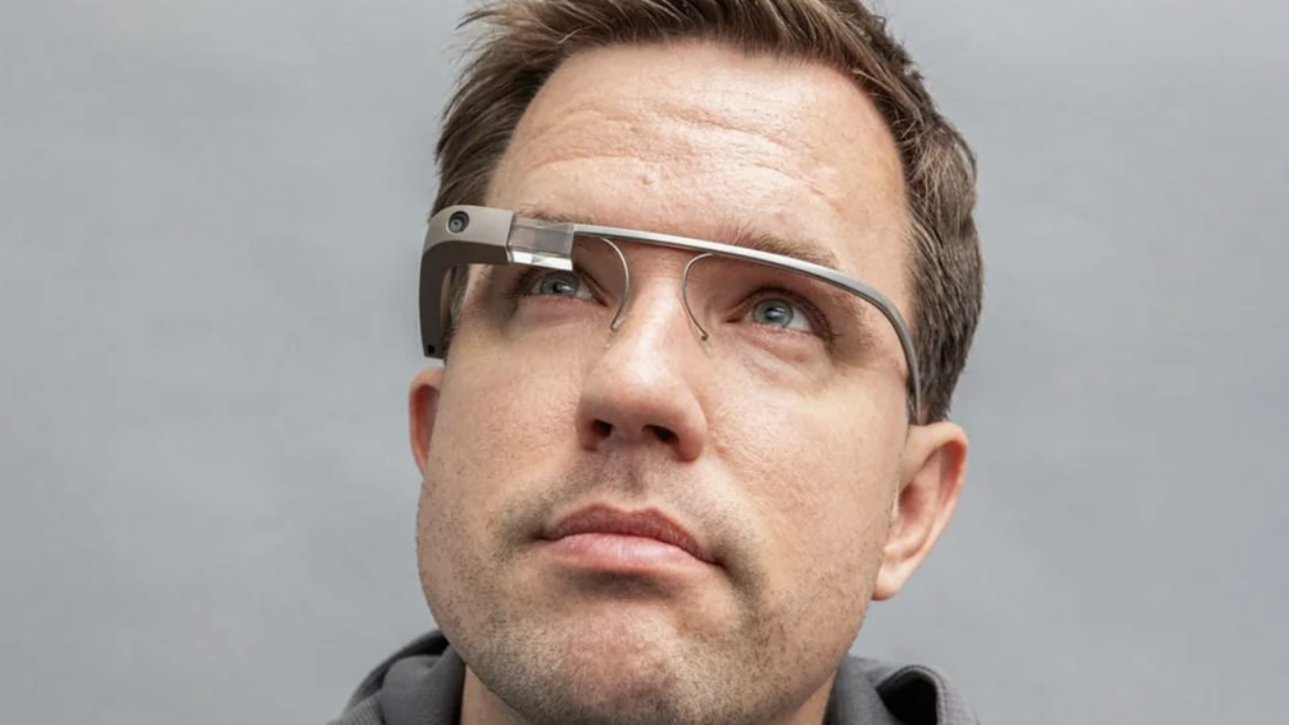 A person wearing smart glasses, one of the latest tech gadgets, looks upward. The background is plain and gray, and the person has short brown hair and a slight stubble.
