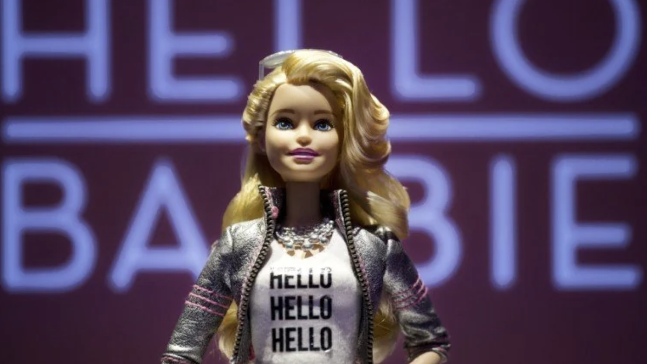 A Barbie doll with long blonde hair stands in front of a pink background with the words “HELLO BARBIE.” She wears a white shirt that says “HELLO” multiple times and a silver jacket, effortlessly blending classic style with the flair of modern tech gadgets.