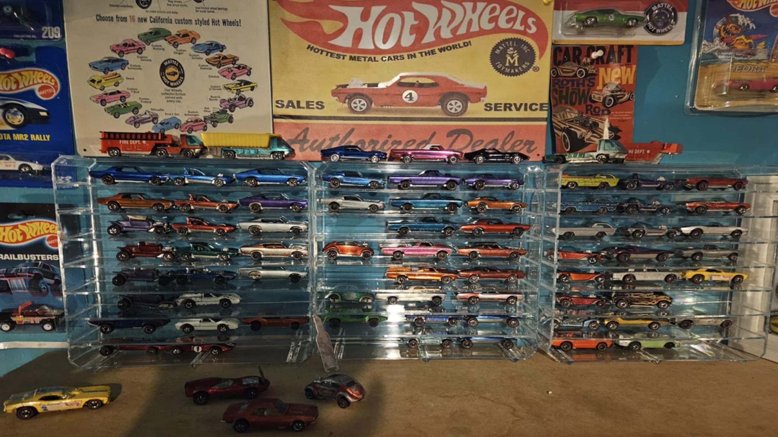 A display of many Hot Wheels toy cars in clear cases, with several cars on the table. Vintage Hot Wheels posters and packaged cars—iconic childhood items—are visible on the wall behind the display.