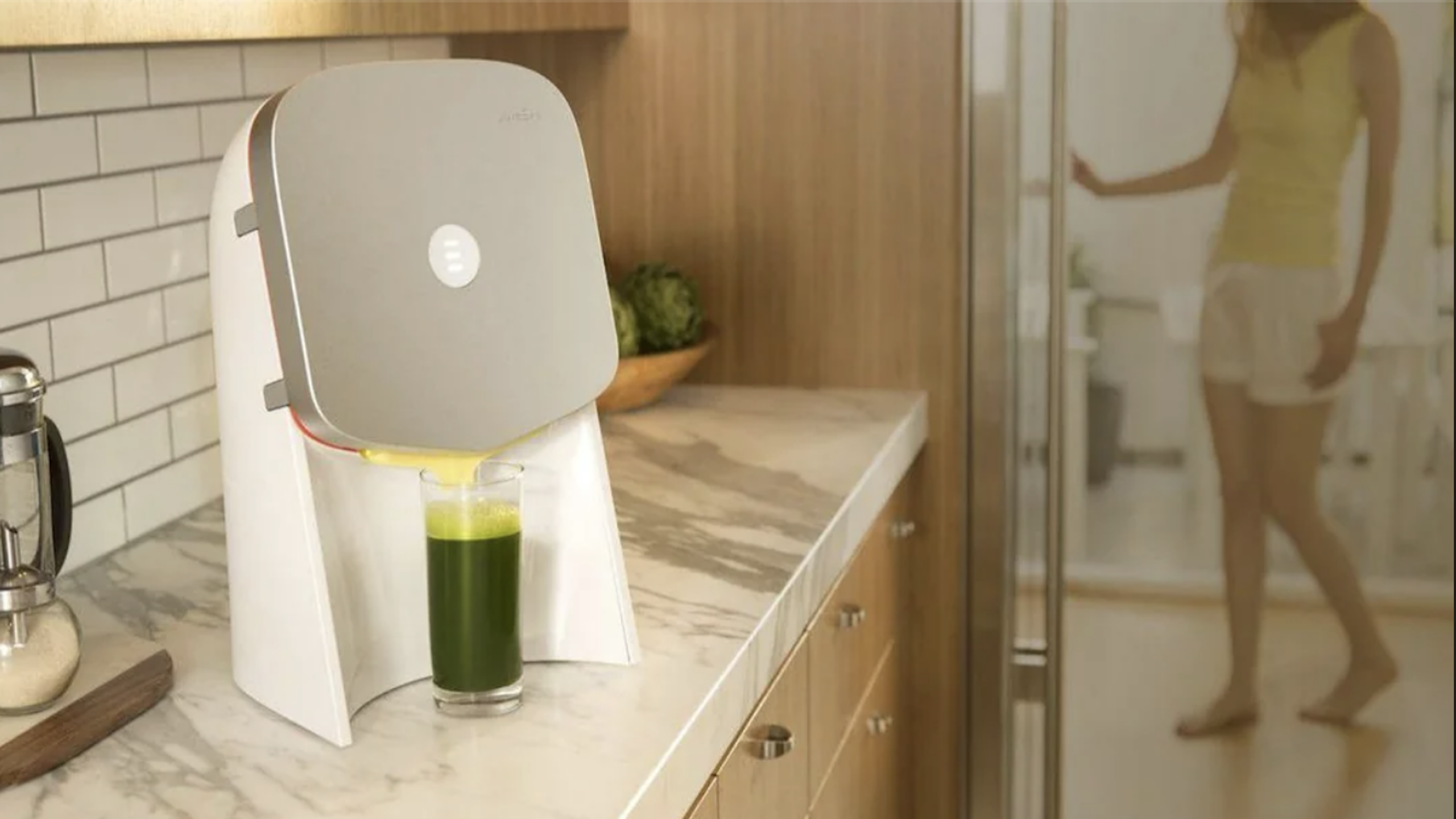 A countertop juice maker, one of the latest tech gadgets, pours green juice into a glass in a modern kitchen. A woman in white shorts and a tank top stands in the background near a refrigerator.