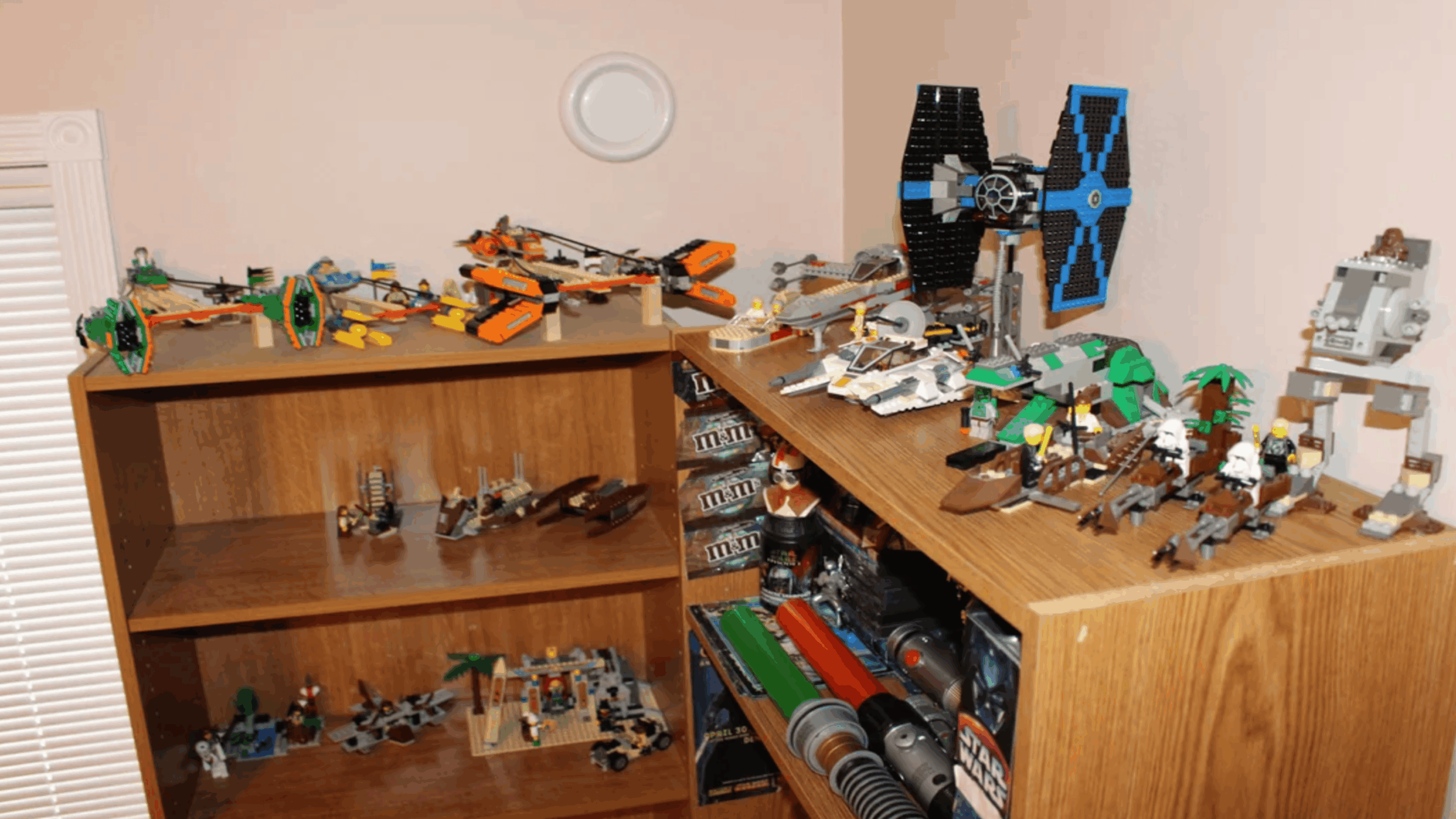 Two wooden shelves filled with assembled Lego Star Wars sets, including ships, walkers, and small vehicles—cherished childhood items—are placed in the corner of a room with beige walls.