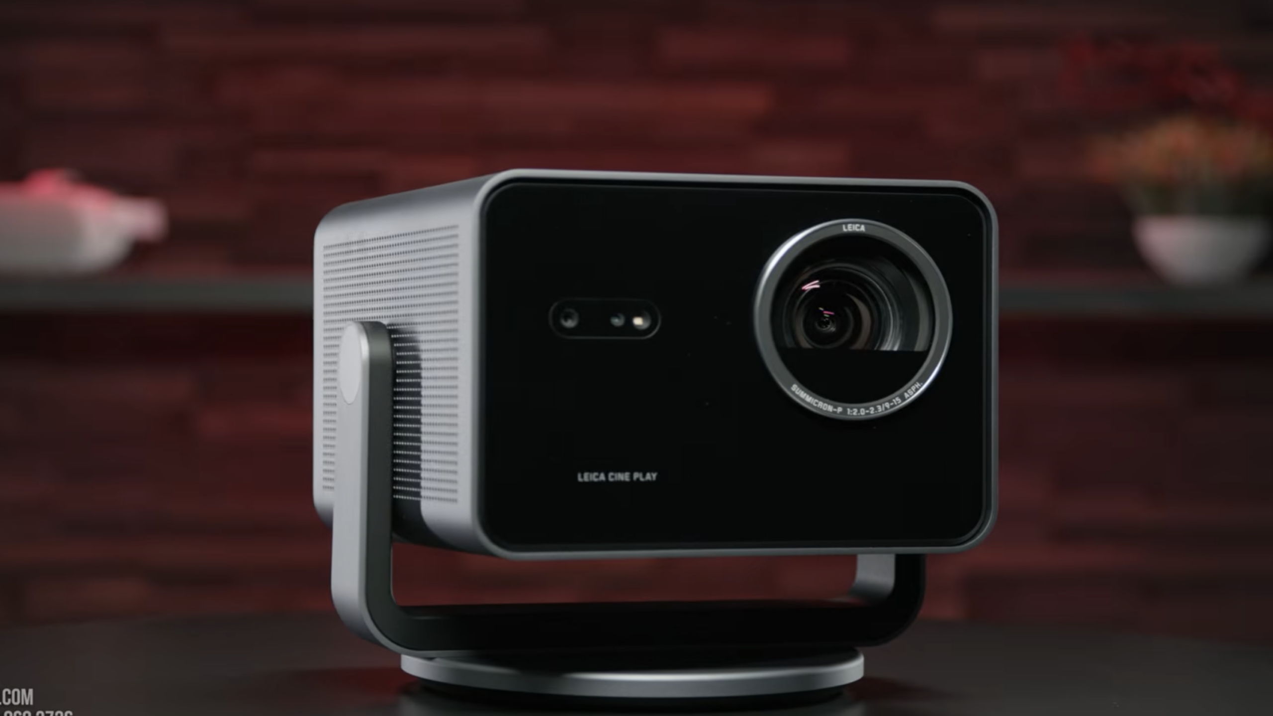 A black Leica Cine 1 projector, ideal for showcasing gaming products, sits on a table against a blurred red and black background, with its lens and control buttons clearly visible on the front panel.