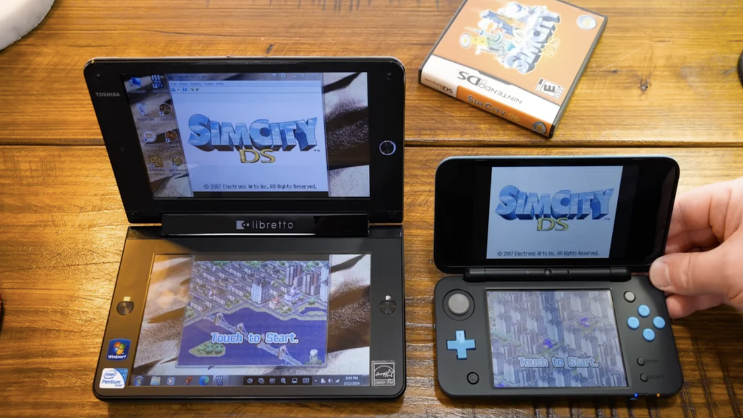 Two tech gadgets display SimCity DS: a black Toshiba Libretto on the left and a blue-and-black Nintendo 2DS XL on the right. A game case with the SimCity art is on the wooden table in the background.