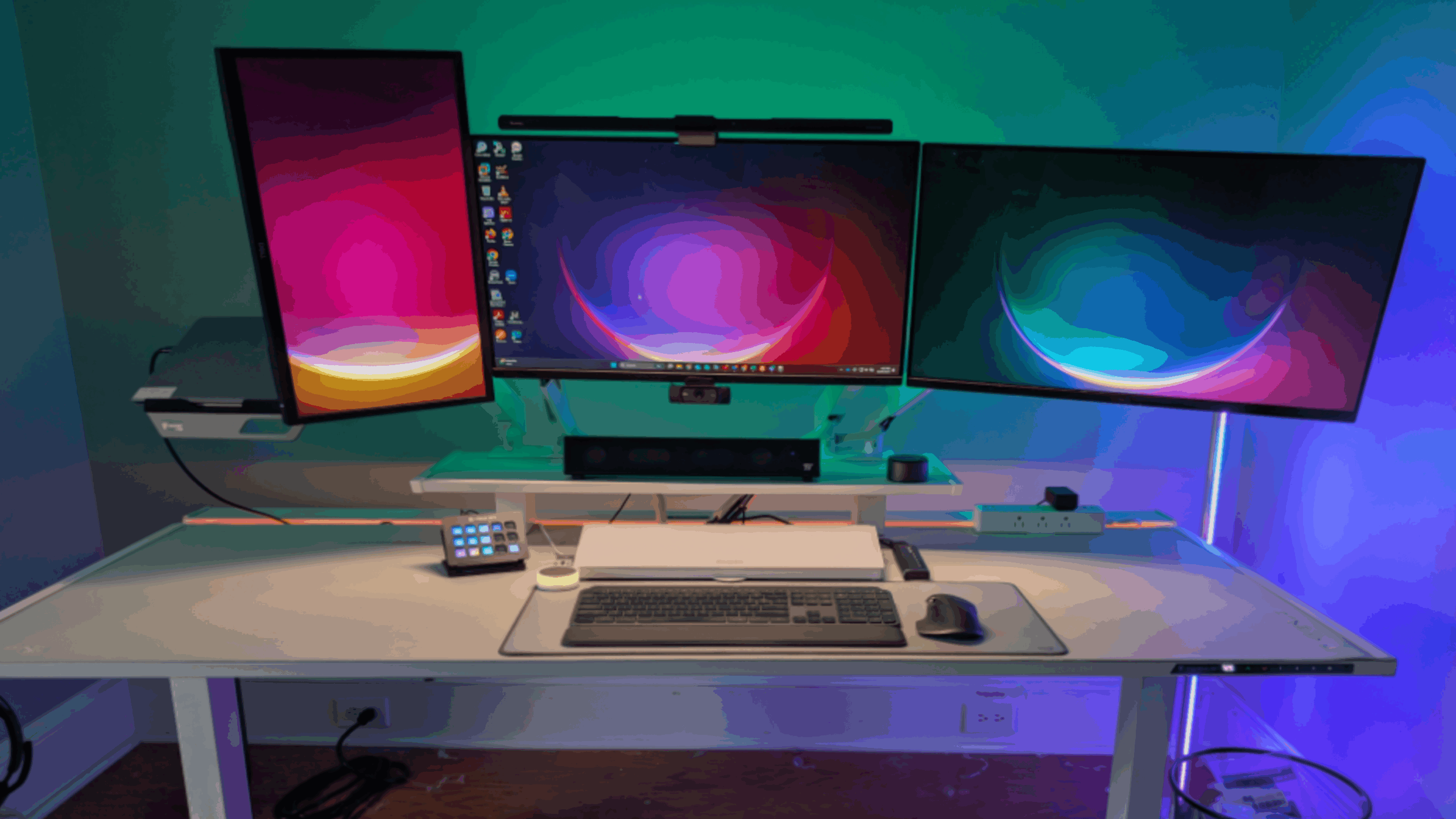 A modern desk setup featuring gaming products, with three colorful monitors—one vertical—a keyboard, mouse, stream deck, speakers, and a drink on a white desk, all illuminated by blue and green LED lights.