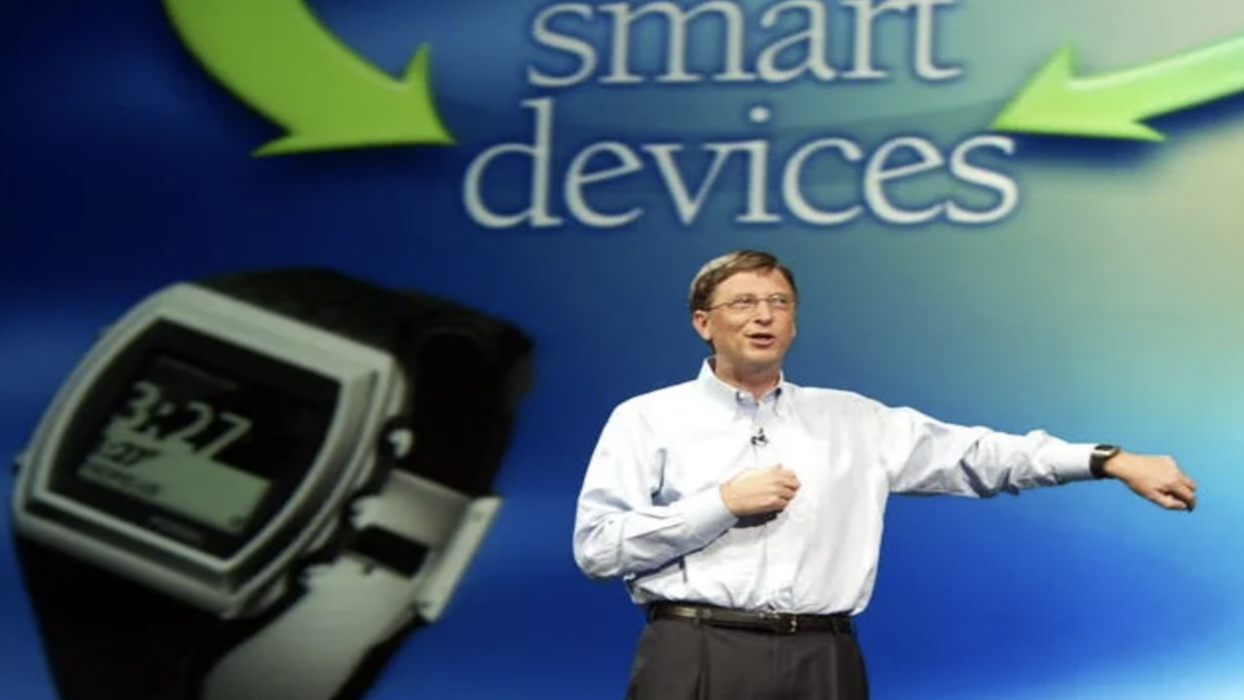 A man in glasses gestures while standing in front of a screen displaying a large smartwatch and the words “smart devices,” highlighting the latest innovations in tech gadgets.