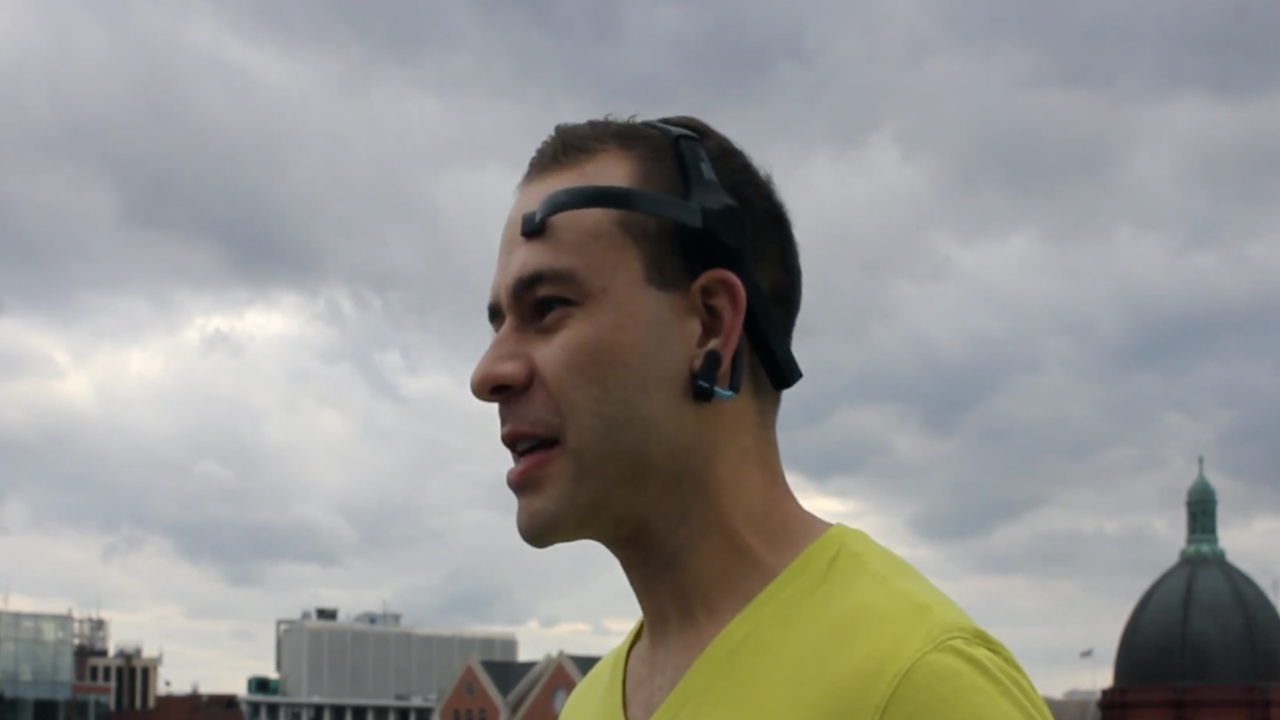 A person wearing a head-mounted tech gadget stands outdoors against a cloudy sky, with some city buildings and a dome visible in the background.