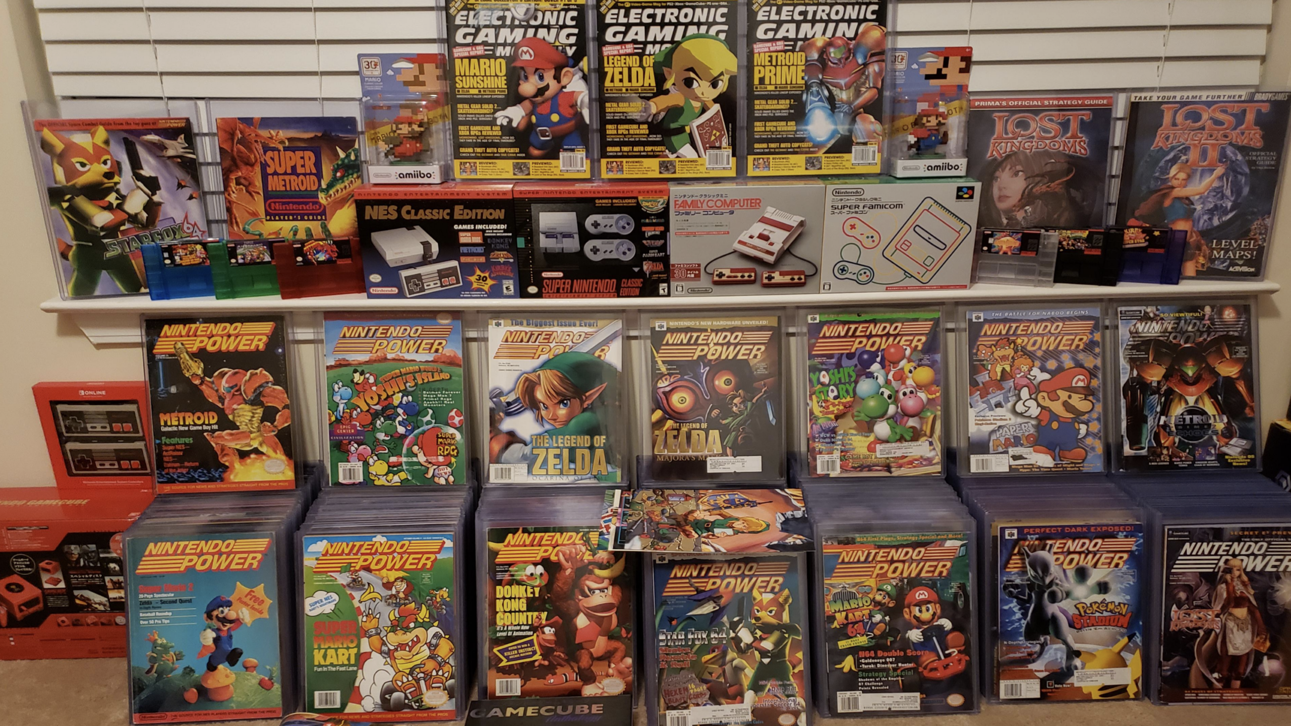 A collection of Nintendo Power and gaming magazines, childhood items like vintage game boxes, and consoles neatly displayed on shelves, featuring Super Mario, Zelda, Metroid, GameCube, and classic NES memorabilia.