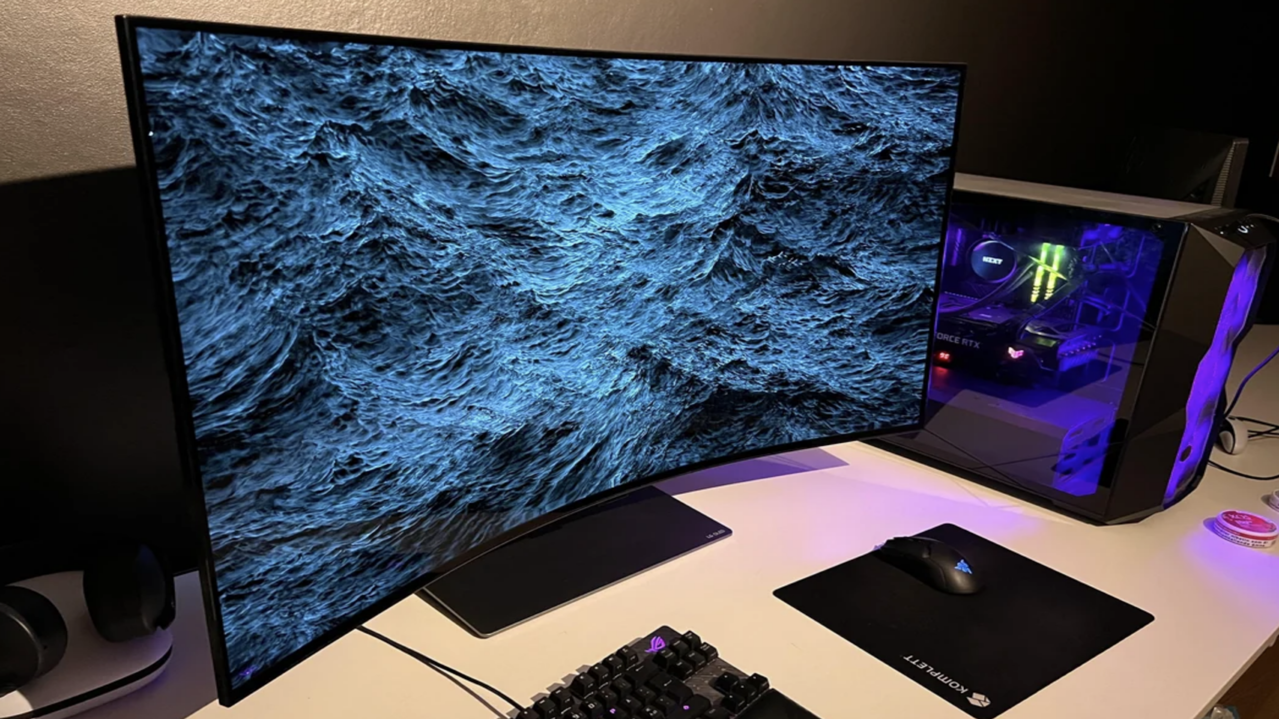 A curved monitor displays a detailed ocean wave wallpaper on a desk, next to gaming products such as a gaming keyboard, mouse, black mousepad, headphones, and a desktop computer with RGB lighting.
