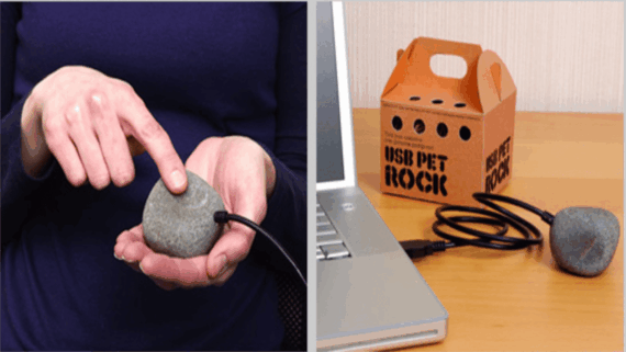A person touches a USB-connected rock near a laptop; next to it is a small box labeled &ldquo;USB Pet Rock,&rdquo; with the quirky tech gadget and cable on a desk.