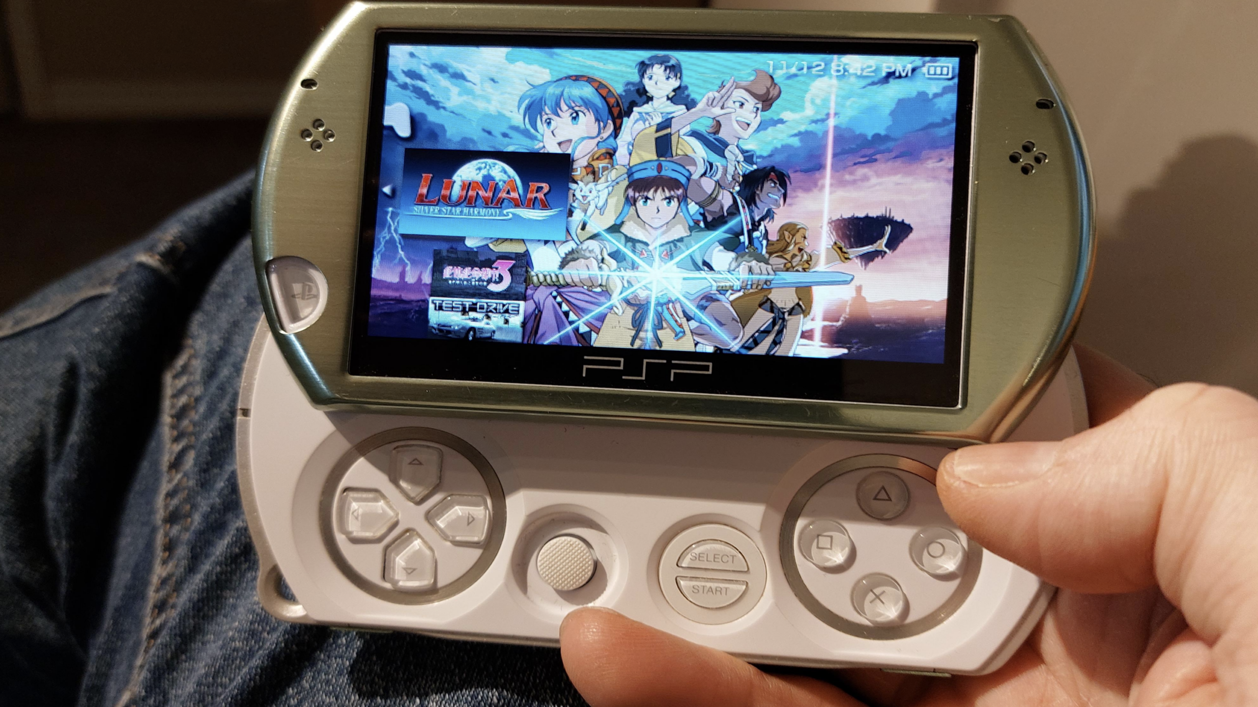 A person holds a white PSP Go, Sony’s compact PlayStation handheld, displaying the game “Lunar: Silver Star Harmony” on its screen. The device rests on their jeans, and their right thumb hovers near the control buttons.