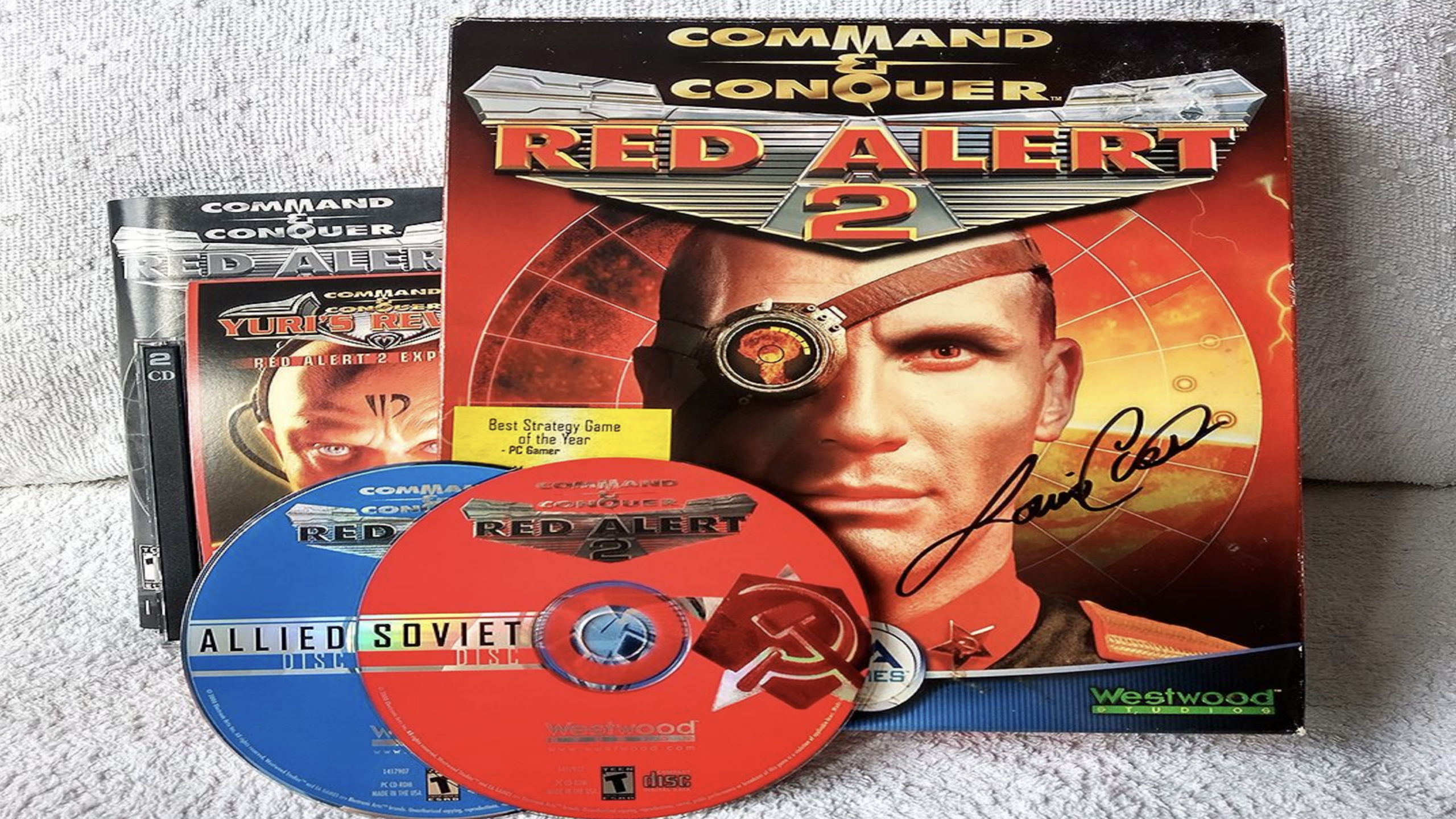 Command & Conquer: Red Alert 2 game box with two distinct game discs labeled “Allied” and “Soviet,” accompanied by the Yuri’s Revenge expansion pack—all displayed on a light surface—featuring a signed cover that showcases a stern man in uniform.