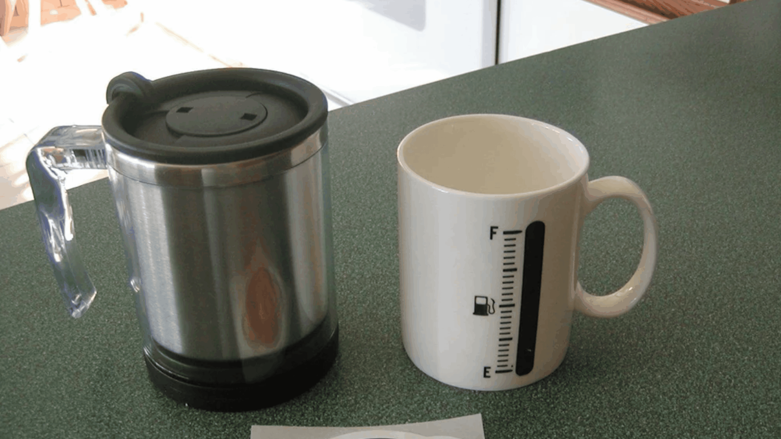 A stainless steel travel mug with a black lid sits next to a white ceramic mug featuring a fuel gauge graphic on a green countertop, making it an ideal setup for tech gadgets enthusiasts looking to stay caffeinated.