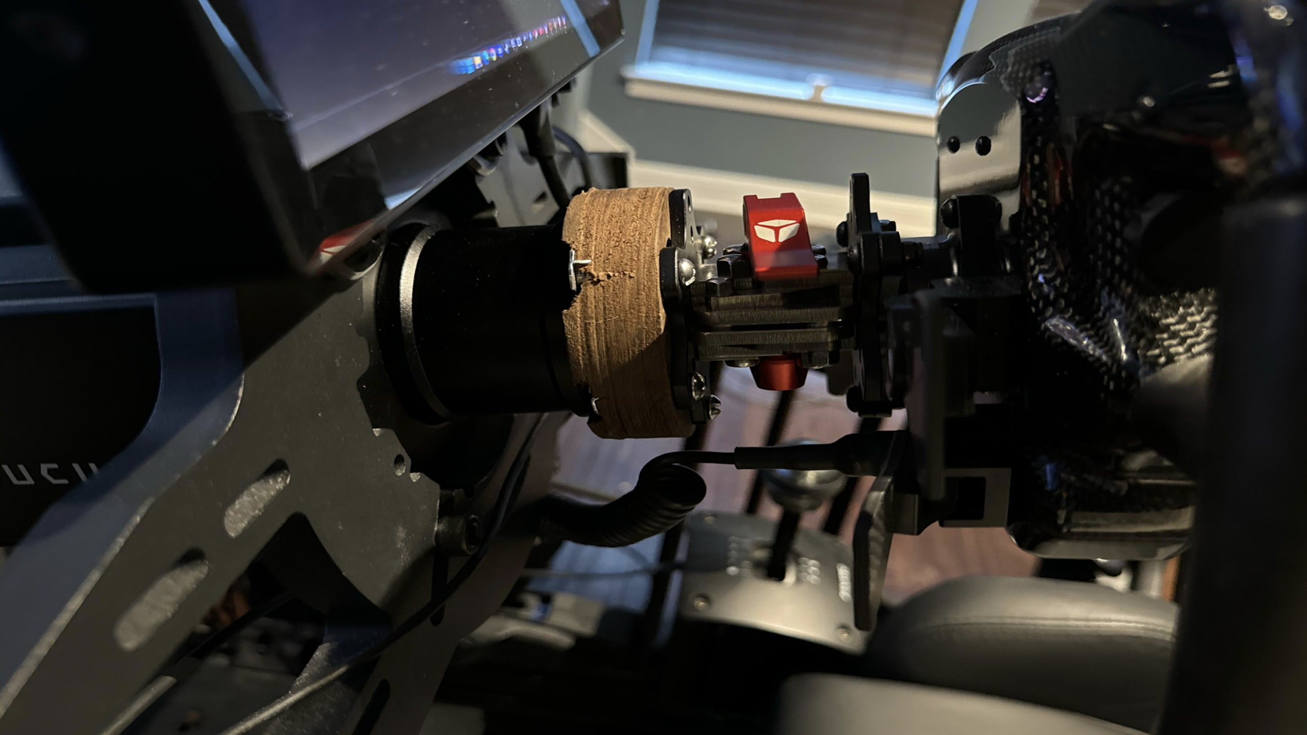 A close-up of a sim racing wheel setup, featuring top-tier gaming products, shows a monitor, a steering wheel with a brown grip, a red quick-release, and seat, all in a dim room with light from a window in the background.