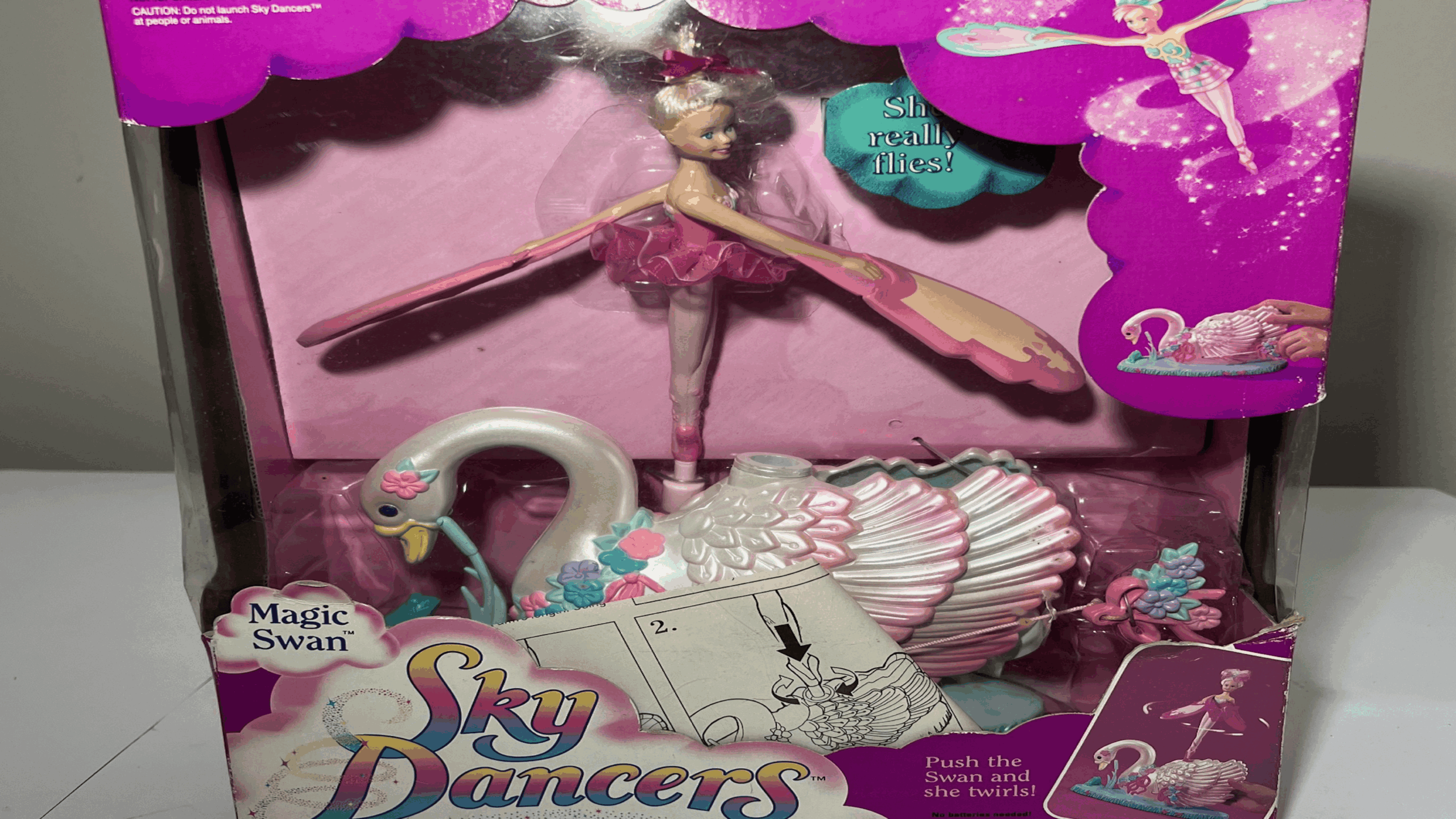 Sky Dancers Magic Swan toy in packaging, featuring a ballerina doll with wings and a white swan base with pink and blue accents. Reminiscent of beloved childhood items, the box is decorated with instructions and bright graphics that capture the magic and wonder of playtime.