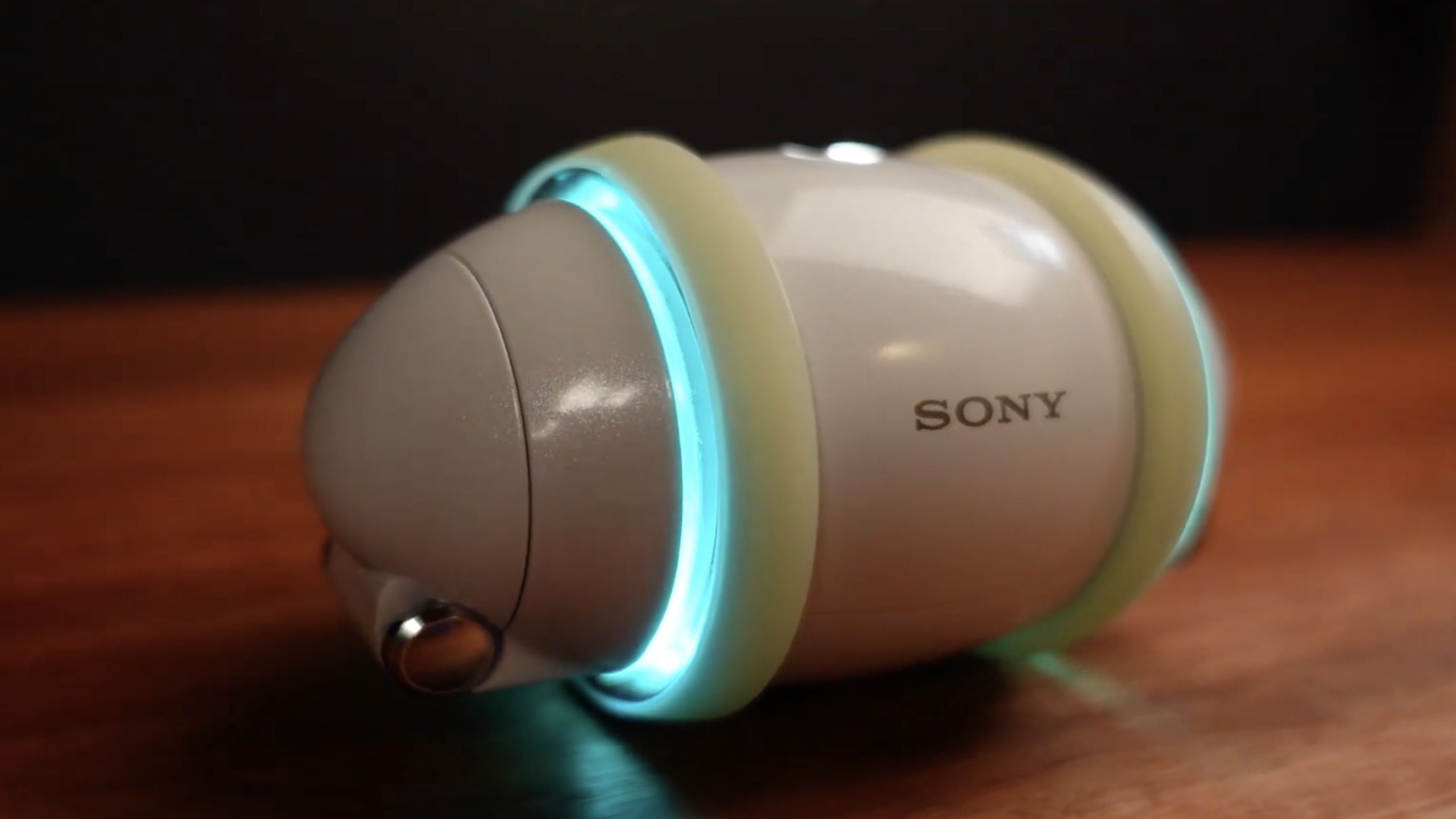 A small, egg-shaped white robot with glowing blue lights and the SONY logo sits on a wooden surface. The robot has rounded edges and metallic details, making it a sleek addition to any modern collection of tech gadgets.