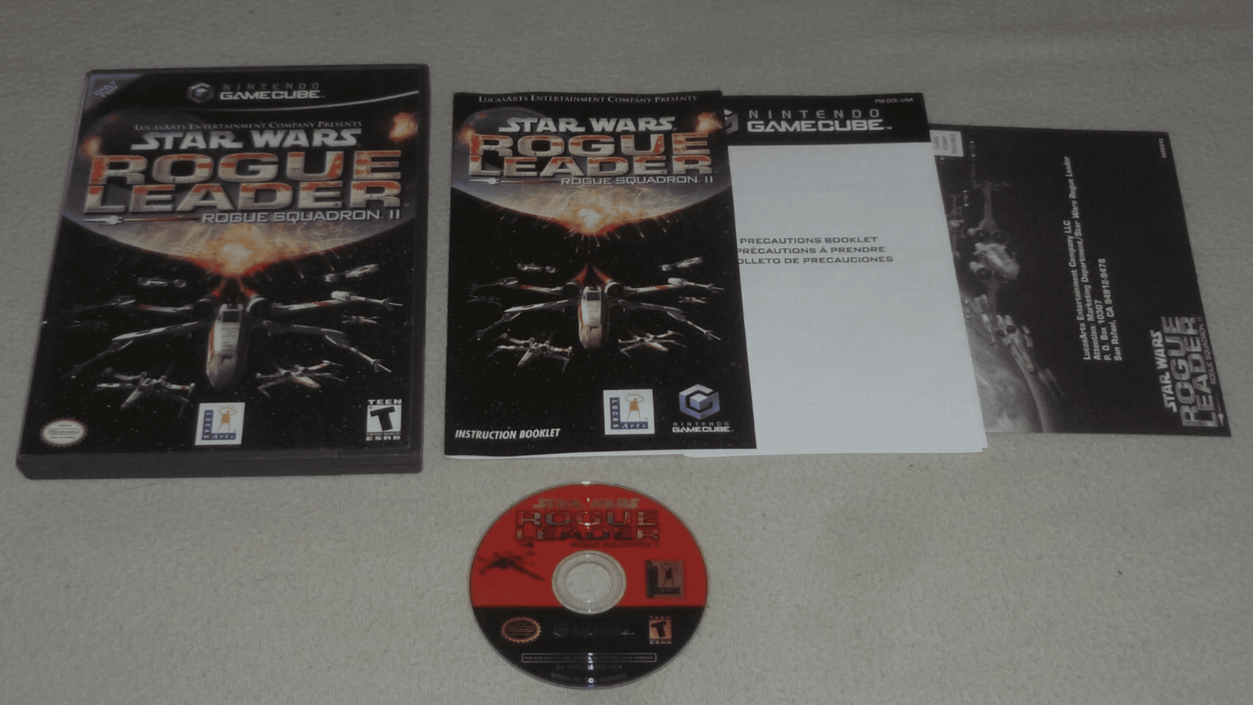 Nintendo GameCube “Star Wars Rogue Leader: Rogue Squadron II” game case, instruction booklet, precaution booklet, and Game Discs are displayed on a light surface.