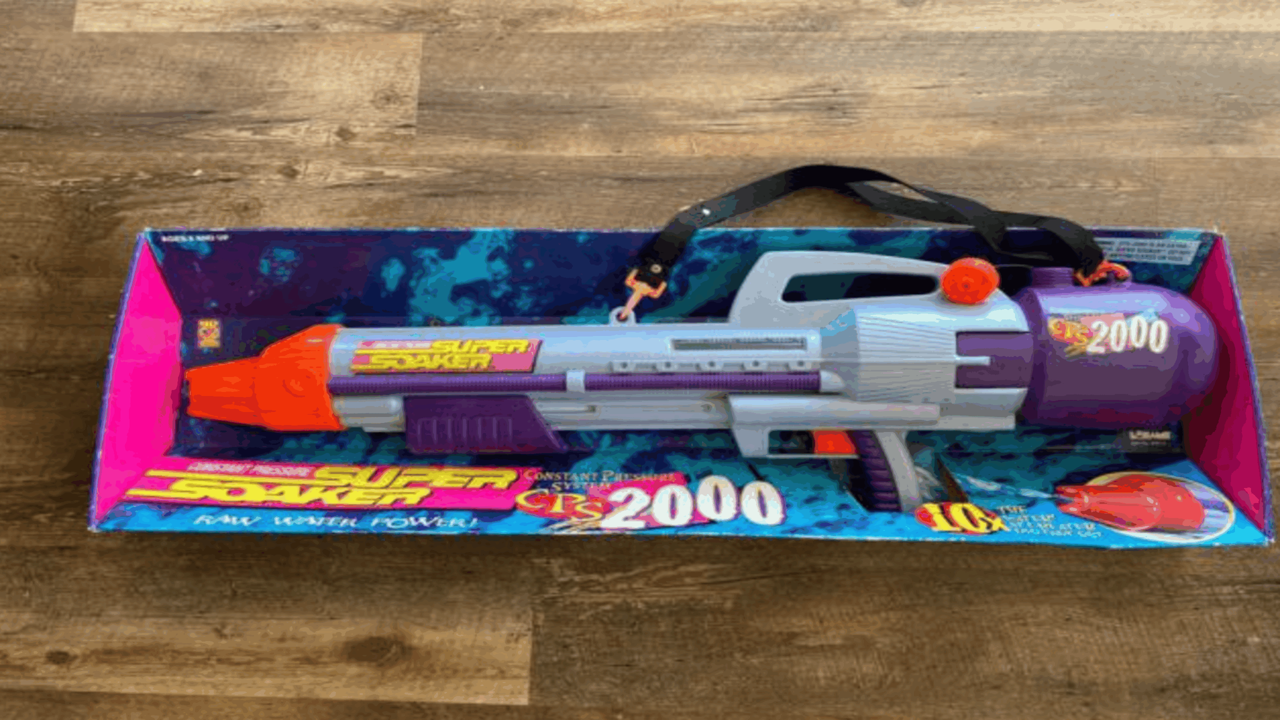 A classic example of coveted childhood items, this Super Soaker CPS 2000 water gun sits in its original packaging on a wood floor. The toy is purple, gray, and orange, with a carrying strap and the iconic CPS 2000 branding visible on the box.