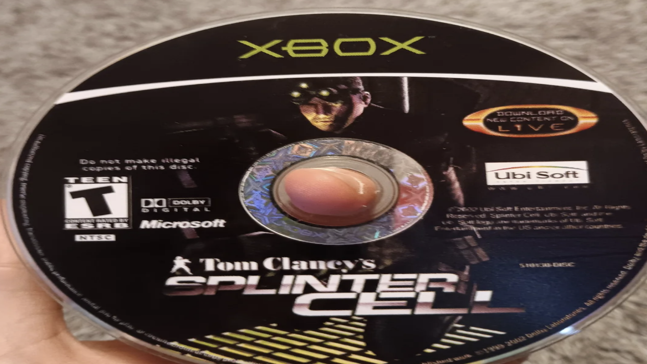 An Xbox Game Disc for “Tom Clancy’s Splinter Cell” shows a character in night vision goggles, with logos for Ubisoft, Dolby Digital, and Xbox across the front. This is one of the classic Game Discs that defined stealth-action gaming.