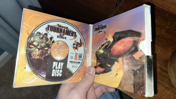 A hand holds an open Unreal Tournament 2004 game case, revealing the Play Disc on the left and iconic Game Discs artwork featuring a vehicle explosion scene on the right. The case is held over a wooden surface.