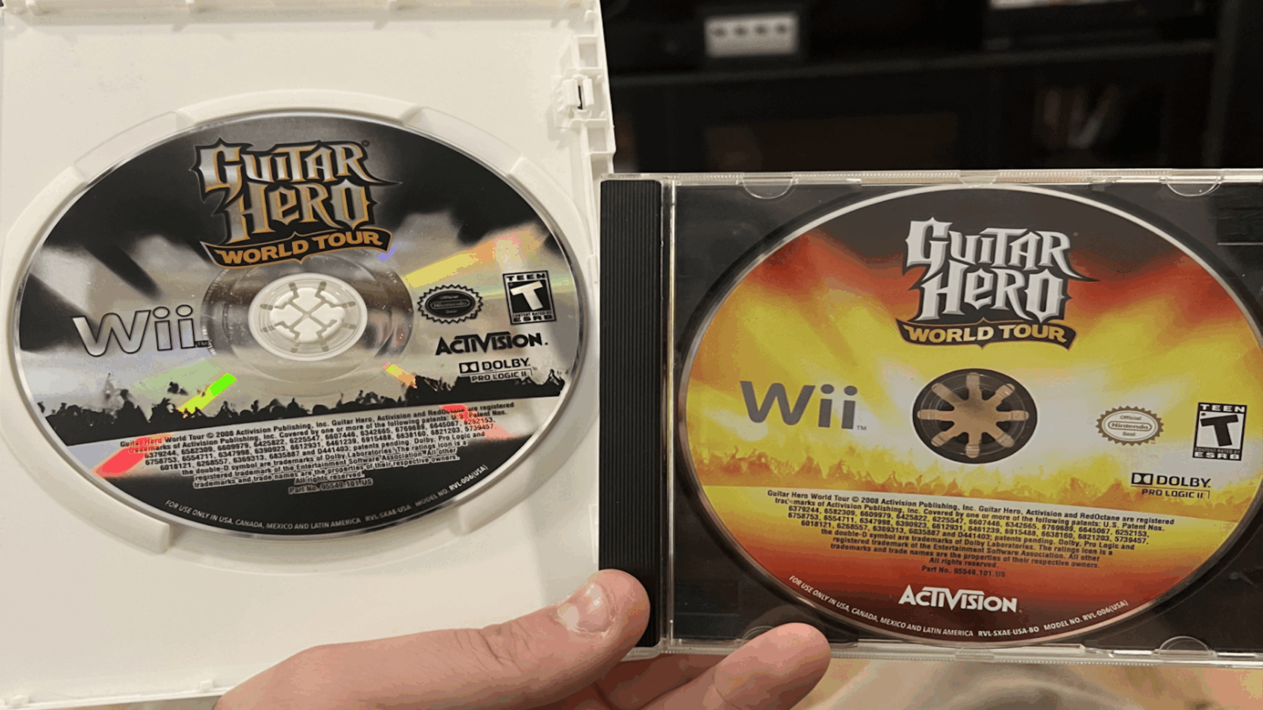 Two Guitar Hero World Tour Wii game discs are shown; one Game Disc is in a white Wii case, the other is in a black CD case. Both Game Discs feature the same logo but have different designs. A hand holds the cases.