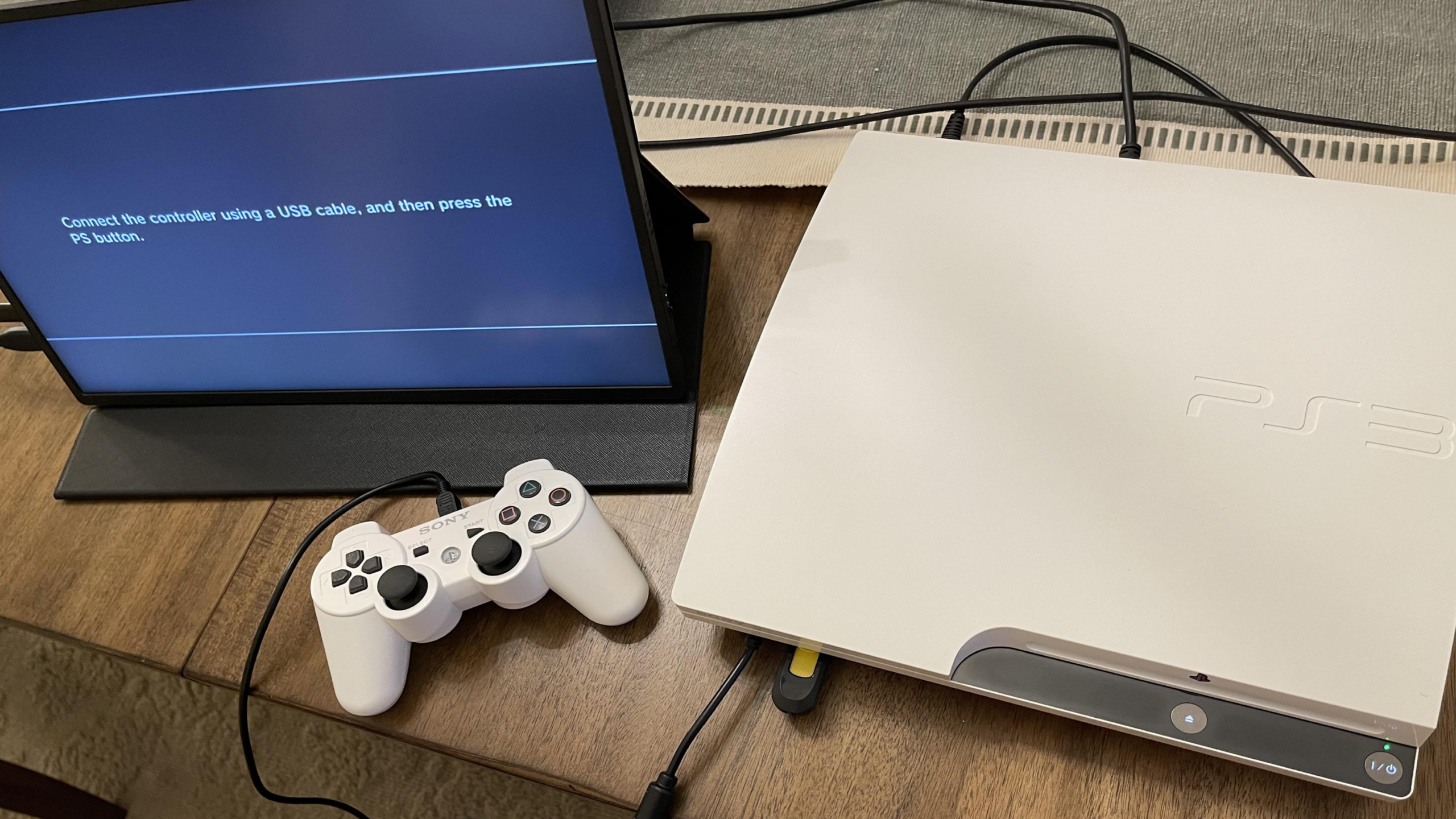 A white PlayStation 3 and white controller sit on a desk next to a monitor displaying instructions to connect a PlayStation controller with a USB cable and press the PS button.